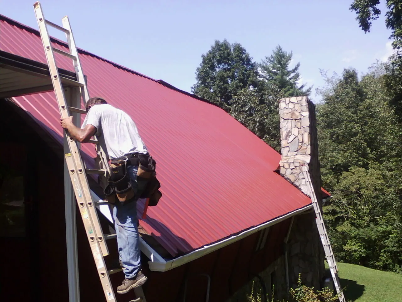 Roofer on a ladder beside a red metal roof and stone chimney — Soffit & Fascia Repair by Roofing Brothers in Tewksbury